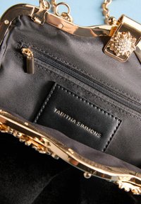 Interiorul unei genți de mână din satin negru cu accesorii din aur, un compartiment cu fermoar și un eticheta cu inscripția "TABITHA SIMMONS", având un închizător decorativ.