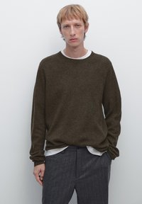 Brun strikket sweater med lange ærmer og rund hals, båret over en hvid skjorte med krave, parret med mørke smalstribede bukser.