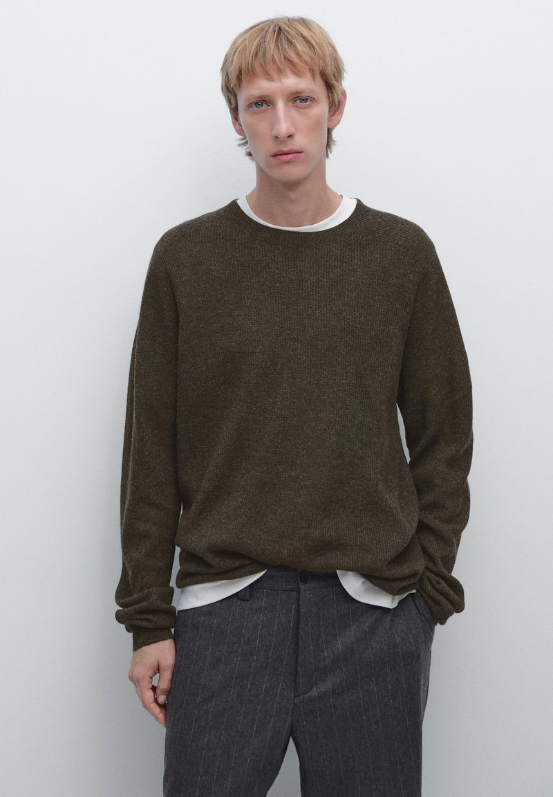 Brun strikket sweater med lange ærmer og rund hals, båret over en hvid skjorte med krave, parret med mørke smalstribede bukser.
