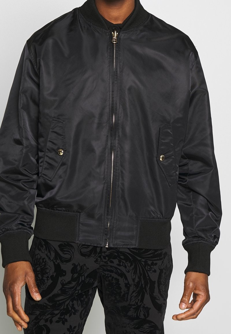 Homme portant une veste bomber noire zippée avec des poches boutonnées et un pantalon noir à motifs, debout devant un fond uni.