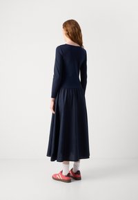 Tummansininen pitkähihainen mekko, jossa on tiukka yläosa ja lentoava helma. Mallilla on päällään vaaleanpunaiset tennarit, joissa on valkoisia yksityiskohtia, sekä vaaleanharmaat sukat.
