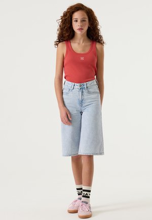 Rotes Tanktop mit kleinem Logo, hochtaillierte hellblaue Jeans-Culottes, pinke Sneakers und schwarz-weiß gestreifte Socken. Welliges braunes Haar.