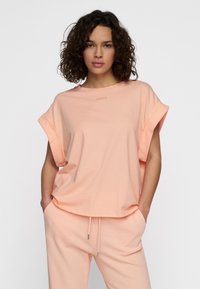 Camiseta de color melocotón, de manga corta con puños enrollados, hecha de una tela suave; presenta un logo sutil en el cuello. Combinada con joggers a juego.
