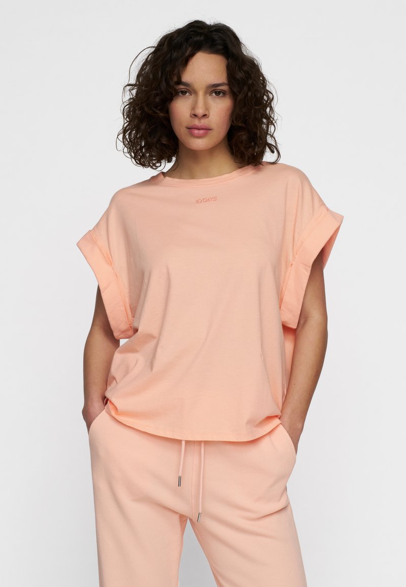 Camiseta de color melocotón, de manga corta con puños enrollados, hecha de una tela suave; presenta un logo sutil en el cuello. Combinada con joggers a juego.
