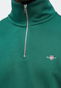 Vihreä pusero, jossa korkea kaulus, puolivetoketjullinen metallisuljenta ja kirjailtu logo. Pehmeä rakenne ja muotoiltu istuvuus.