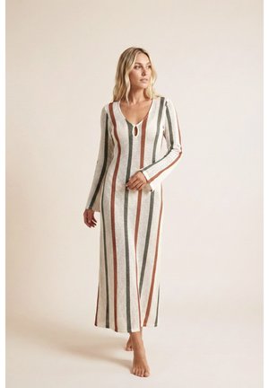 Mujer de pie descalza con un vestido largo de punto con rayas verticales verdes, óxido y crema y escote en forma de ojo de cerradura.