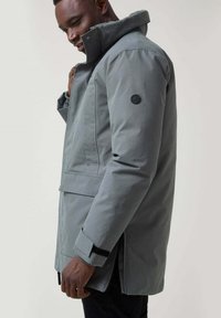 Chaqueta impermeable gris con alto cuello, bolsillos laterales y parche de logo en la manga. Presenta un corte recto y puños ajustables.