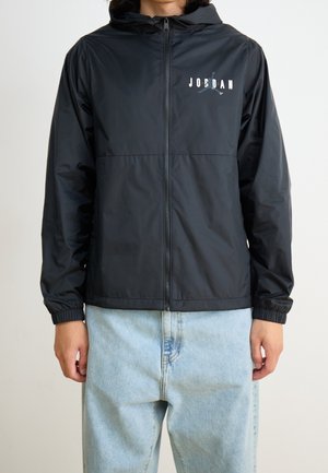 Person iført en sort Jordan lynlås-hættetrøje med hætte og elastiske manchetter, kombineret med lyseblå denimjeans.