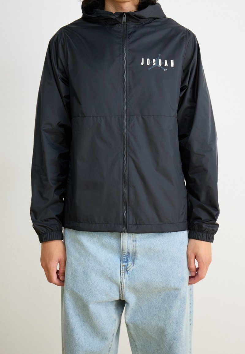 Personne portant un hoodie noir Jordan zippé avec capuche et poignets élastiques, associé à un jean denim bleu clair.