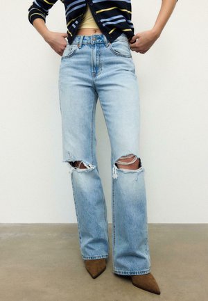 Osoba oblečená v světle modrých roztrhaných džínách s ošuntělými dírami na kolenou a hnědých špičatých botách, stojící proti jednoduché stěně a podlaze.