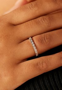 Silver band ring featuring multiple small, round clear stones set in a row, displayed on a hand with a textured, tanned skin tone.