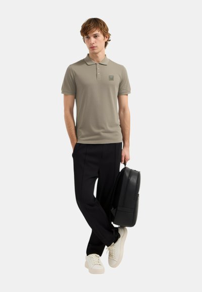 Olivno zelena polo majica s ovratnikom i tremi gumbi, uparena s crnim hlačama i bijelim tenisicama, drži crni ruksak.