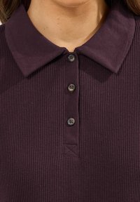 Diep paarse geribde polo met een zachte kraag en vier ronde knopen met bijpassende draad. De stof heeft een gestructureerde uitstraling.