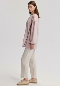 Lichtroze blouse met lange mouwen en puffmouwen, voorzien van zwarte knoopdetails, gecombineerd met crème rechte broek en beige veterschoenen.