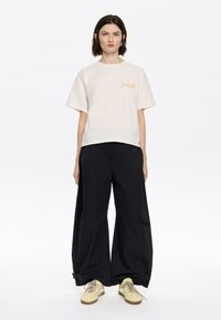 Crème kleurige crop top met korte mouwen en geborduurde gouden tekst, gecombineerd met losse zwarte broeken en gele sneakers. Eenvoudig, minimalistisch ontwerp.