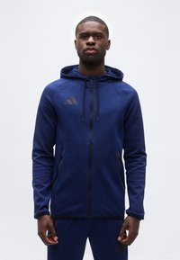 Άνδρας που φοράει μπλε σκούρο φούτερ με φερμουάρ και ταιριαστό παντελόνι με μαύρες λεπτομέρειες, στέκεται μπροστά σε ένα απλό λευκό φόντο.