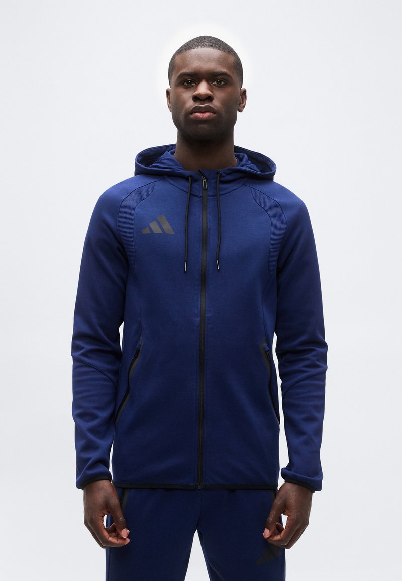 Άνδρας που φοράει μπλε σκούρο φούτερ με φερμουάρ και ταιριαστό παντελόνι με μαύρες λεπτομέρειες, στέκεται μπροστά σε ένα απλό λευκό φόντο.