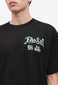 Κοντινό πλάνο ενός ατόμου που φοράει μαύρο t-shirt Diesel με λευκό κείμενο που γράφει "Diesel since 1978 fine quality" στο στήθος.