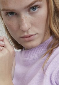Nahaufnahme einer Person, die einen hell-lila gestrickten Pullover mit einem gerippten Halsauschnitt trägt. Sichtbare Sommersprossen und helles Haar.