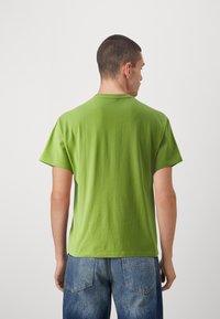 Zelena bombažna majica s krojem okrog vratu in kratkimi rokavi, videna zadaj. Oblikovana z modrimi kavbojkami, kar prikazuje sproščen fit.