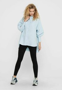 Camicia oversized azzurro chiaro con bottoni e maniche lunghe a sbuffo, abbinata a leggings neri e sneakers blu. Design semplice e look casual.
