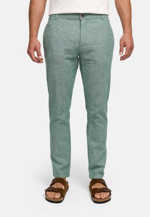Hombre con pantalones ajustados de color verde claro y sandalias marrones con doble tira, de pie contra un fondo blanco.