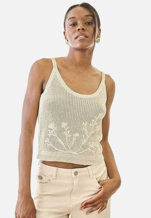 Mujer joven con un top de punto color crema con patrón floral y pantalones beige claro, posando con una mano en el bolsillo contra un fondo blanco.