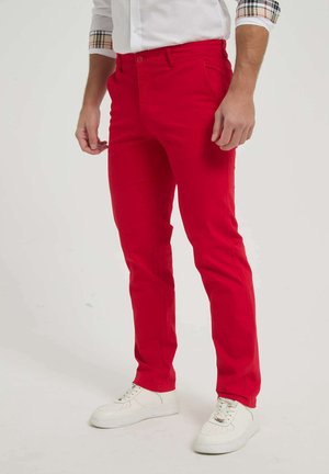 Homme portant un pantalon rouge vif, des baskets blanches et une chemise blanche à boutons avec des manches retroussées à motifs à carreaux, debout sur un fond uni.