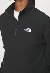 Personne portant une polaire noire à fermeture éclair quart de zip avec le logo blanc "The North Face" sur la poitrine gauche, par-dessus une chemise blanche.