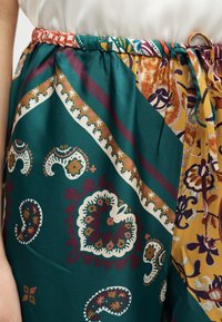 Zone de la taille et des hanches d'une jupe à motifs avec des designs cachemire et floraux en sarcelle, moutarde, blanc et bordeaux, taille froncée et fine ceinture avec anneau métallique.