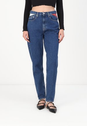 Osoba ubrana w niebieskie, wysokie w talii dżinsy z kolorowymi detalami kieszeni, czarny crop top z długim rękawem i czarne, szpiczaste baleriny.