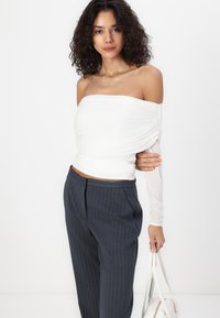 White ruched off-shoulder top with long sheer sleeve, paired with dark pinstriped pants. The model holds a white handbag.
