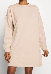 Robe-sweat à manches longues, col rond, de couleur beige. Fabriquée en tissu doux avec une coupe décontractée et des poignets côtelés, tombant au-dessus de la mi-cuisse.