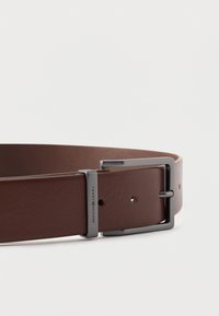 Brunt læderbælte med en sølvfarvet metal spænde. Bæltet har en glat tekstur med synlige syninger og et logo på spænden.