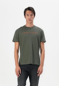 Olijf groene katoenen T-shirt met korte mouwen, ronde halslijn en oranje "TOMMY JEANS" tekst over de borst. Draag het met donkere jeans.