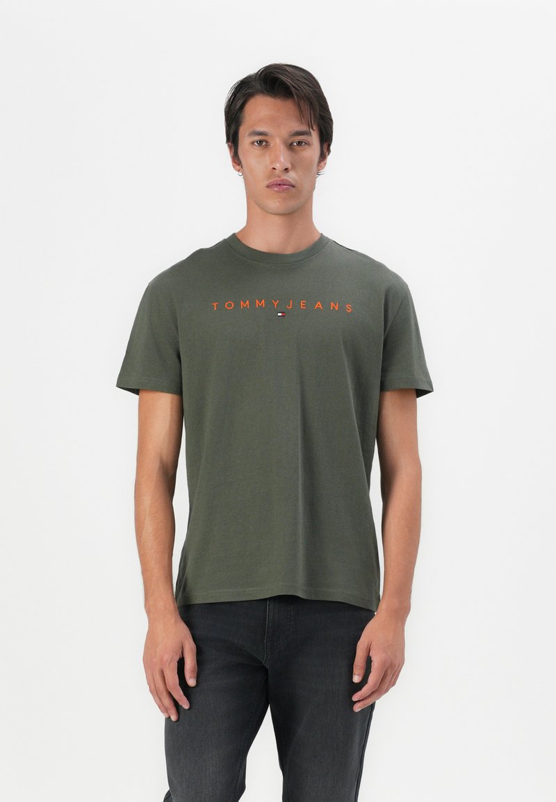 Olijf groene katoenen T-shirt met korte mouwen, ronde halslijn en oranje "TOMMY JEANS" tekst over de borst. Draag het met donkere jeans.