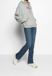 Sweat à capuche gris avec un logo rouge, présentant une coupe ample et un cordon de serrage. Associé à un jean bleu évasé et des baskets claires.