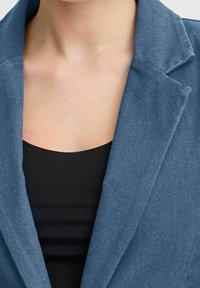 Blå denimblazer med en strukturerad krage, som har en slät svart linne under, vilket visar en lager-på-lager-look.