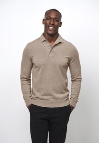 Beige Langarm-Polo-Pullover mit drei Knöpfen, gerippten Bündchen und Saum, kombiniert mit schwarzen Hosen, vor einem weißen Hintergrund stehend.
