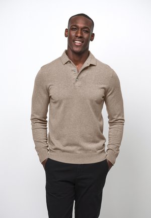 Beige Langarm-Polo-Pullover mit drei Knöpfen, gerippten Bündchen und Saum, kombiniert mit schwarzen Hosen, vor einem weißen Hintergrund stehend.