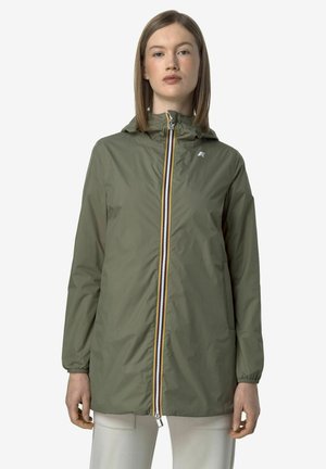 Giovane donna che indossa una giacca antipioggia con cappuccio verde oliva, arricchita da una zip frontale a righe multicolori e pantaloni chiari, in piedi davanti a uno sfondo uniforme.