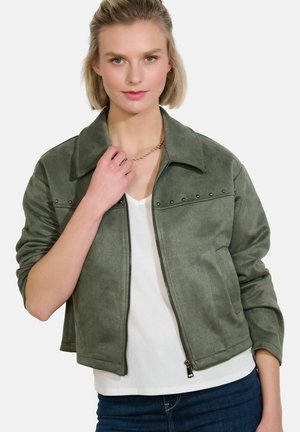 Mladá žena nosiaca krátku olivovo zelenú bundu na zips s detailmi na cvokoch cez biele tričko a modré džínsy, držiaca zlatý retiazkový náhrdelník.