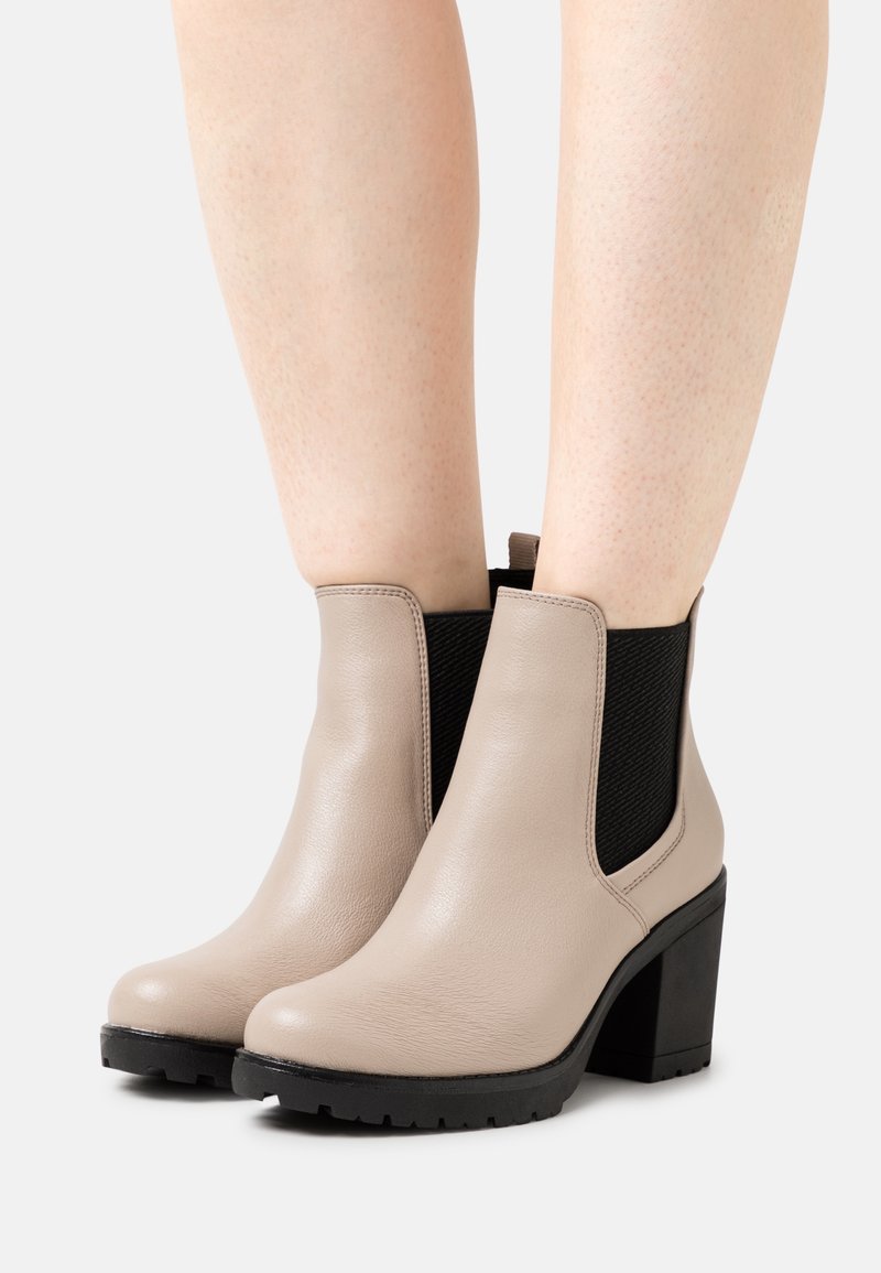 Bottes basses en cuir beige avec côtés élastiques noirs, talons carrés et semelles en caoutchouc texturées. Finition lisse et détails cousus.