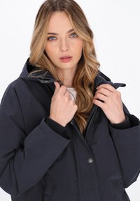 Marineblauwe waterdichte jas met een capuchon, vooraan met rits- en knoopsluiting, ribgebreide manchetten en een gladde textuur.