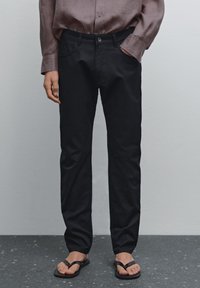 Zwarte broek in een slim fit, met een knoopsluiting, vijf zakken en een soepele katoenmengsel stof. Gecombineerd met zwarte slippers.