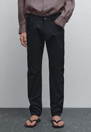 Pantaloni neri aderenti, con chiusura a bottone, cinque tasche e un tessuto morbido in cotone misto. Abbinati a ciabatte nere.