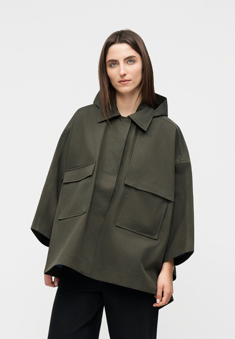 Woman wearing oversized dark green hooded jacket with large front pockets, standing against plain light background.