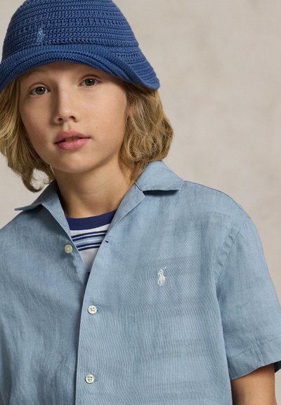 Enfant aux cheveux blonds mi-longs porte un chapeau seau bleu crocheté et une chemise bleu clair boutonnée par-dessus un tee-shirt rayé.