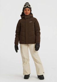 Žena nosi smeđi zimski kaput, bež snježne hlače, crne rukavice, crne čizme, crnu kapu i bijele skijaške naočale na glavi, stojeći pred bijelom pozadinom.