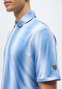 Άνδρας που φοράει ανοιχτό μπλε πόλο πουκάμισο με γιακά και μαύρο έμβλημα με το λογότυπο της Nike για γκολφ στο αριστερό μανίκι, εμφανίζεται από το στήθος και πάνω.